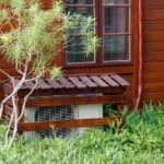 Air conditioning unit partially obscured by grass and a small tree, adjacent to a wooden building, illustrating HVAC installation and maintenance context.