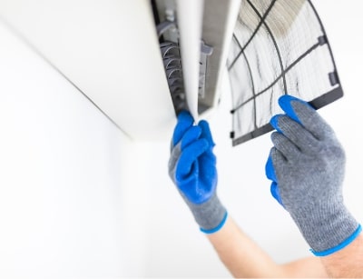 Person wearing blue gloves removing an air conditioning filter for maintenance, emphasizing the importance of HVAC system upkeep before spring.
