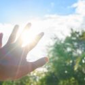 Hand reaching towards sunlight with blue sky and trees in the background, symbolizing summer cooling and HVAC services.