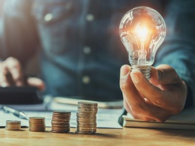 Light bulb illuminating above hand holding it, with stacked coins in the foreground, symbolizing energy efficiency and cost savings related to HVAC systems.