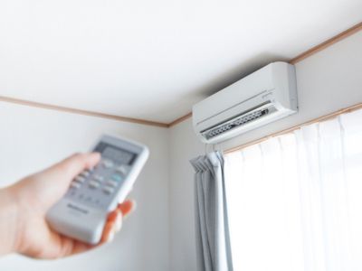 Hand holding remote control aimed at a ductless mini-split air conditioning unit mounted on a wall, illustrating zone control and comfort in HVAC systems.