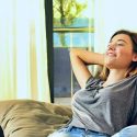 Woman relaxing on a couch, enjoying cool air in a well-lit living room, emphasizing comfort and relaxation in a home environment.