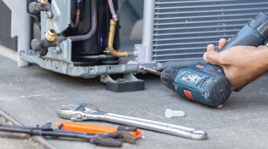 Hand using a power drill on an air conditioning unit, with tools like wrenches and pliers nearby, illustrating air conditioner repair process.